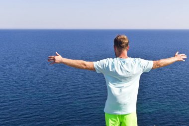 kollarını uzanmış genç bir adam bir uçurumun kenarında duruyor ve denizin muhteşem manzarasına hayran