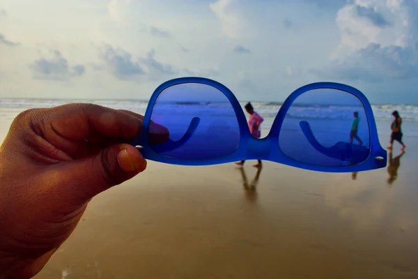Uma mão segurando um vidro de sol na frente do mar.E três pessoas andando — Fotografia de Stock