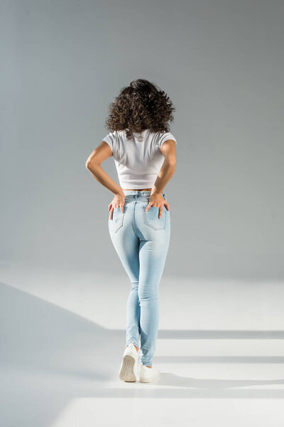 back view of woman standing with hands in pockets in tight blue jeans on grey background