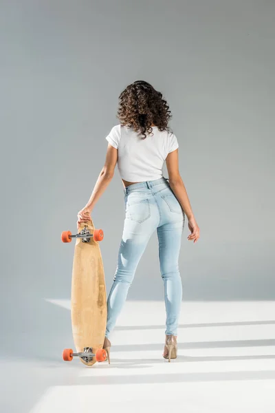 back view of woman with curly hair standing with skateboard on grey background
