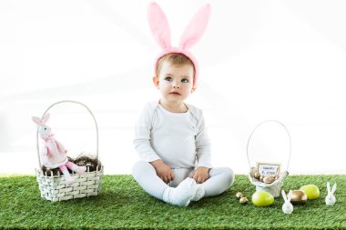 adorable child in bunny ears headband sitting near straw baskets with Easter colorful eggs and decorative rabbits isolated on white clipart