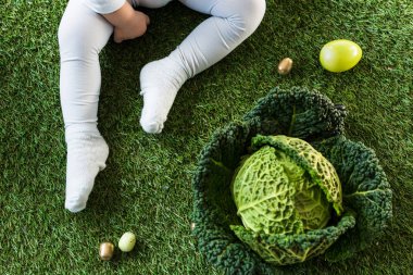 cropped view of baby sitting on green grass near Easter eggs and savoy cabbage clipart