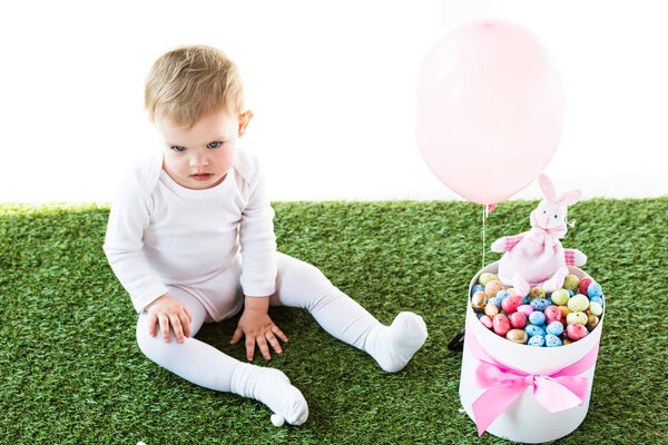 милый ребенок сидит на зеленой траве рядом с коробкой с красочными яйцами перепела, декоративный кролик и воздушный шар изолированы на белом
