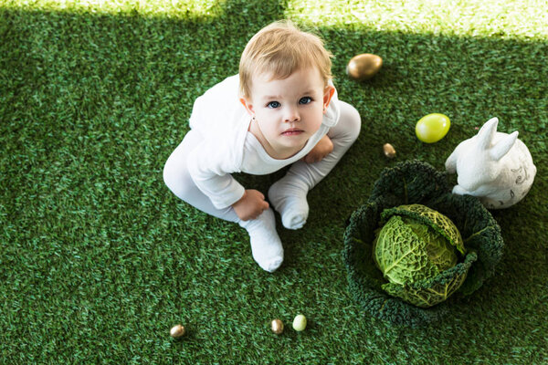 overhead view of cute kid looking at camera while sitting near Easter eggs, decorative rabbit and savoy cabbage