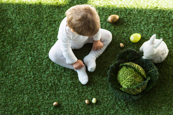 overhead view of cute kid sitting on green grass near Easter eggs, decorative rabbit and savoy cabbage