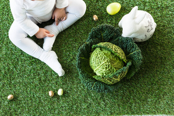 partial view of child sitting on green grass near Easter eggs, decorative rabbit and savoy cabbage