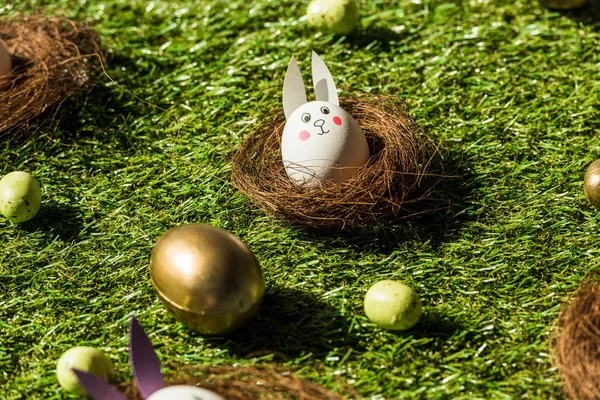 Easter eggs and chicken egg with funny rabbit face and paper ears on green grass