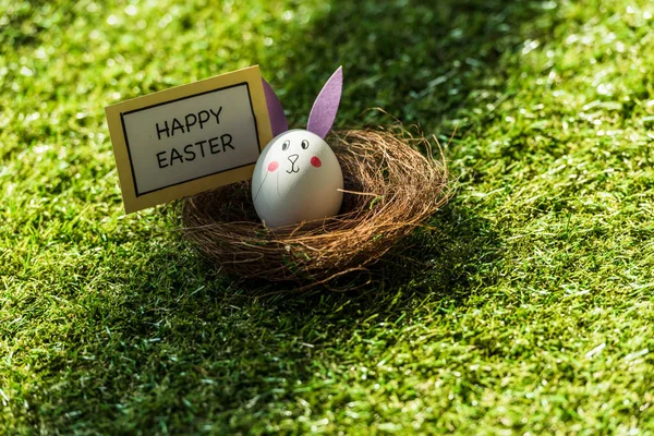 chicken egg with drawn rabbit face and paper ears, and happy Easter card on green grass