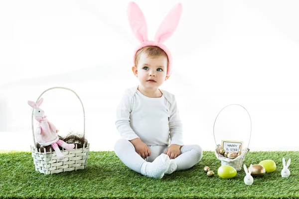 Adorable child in bunny ears headband sitting near straw baskets with Easter colorful eggs and decorative rabbits isolated on white — Stock Image Adorable Child Bunny Ears Headband Sitting Straw Baskets Easter Colorful Royalty Free Stock Photos
