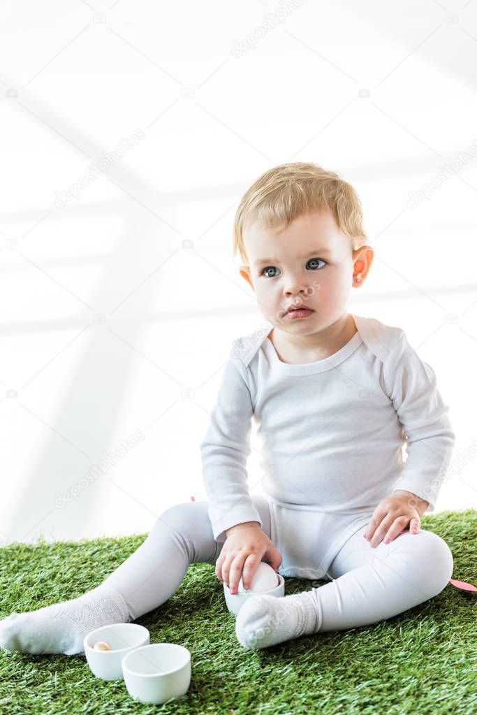 cute baby putting chicken egg into bowl while sitting on green grass isolated on white