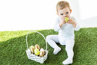 beyaz üzerinde izole Paskalya yumurtaları ile saman sepeti yakın otururken yüz yakın sarı mutfak yumurta tutan sevimli çocuk