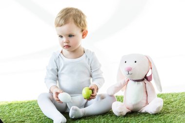 beyaz üzerinde izole oyuncak tavşan yakın yeşil çim üzerinde otururken tavuk yumurtası tutan sevimli çocuk