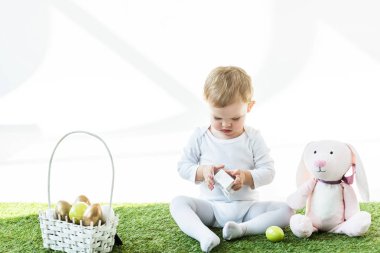 sevimli çocuk beyaz kutu beyaz üzerinde izole Paskalya yumurtaları ile oyuncak tavşan ve saman sepeti yakın yeşil çim üzerinde otururken tutarak