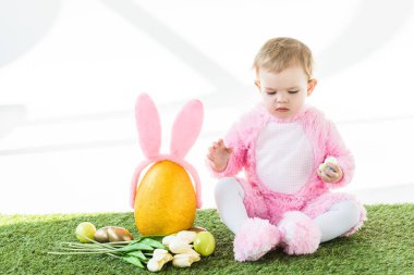 cute kid in pink fluffy costume sitting near yellow ostrich egg with bunny ears headband, colorful Easter eggs and tulips isolated on white clipart