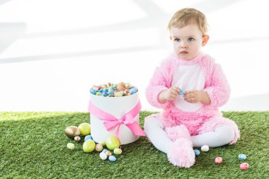 pembe kabarık kostüm sevimli bebek beyaz üzerinde izole renkli Paskalya yumurtaları ile kutu yakın yeşil çim üzerinde oturuyor