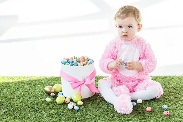 adorable baby in pink fluffy costume sitting on green grass near box with colorful Easter eggs isolated on white