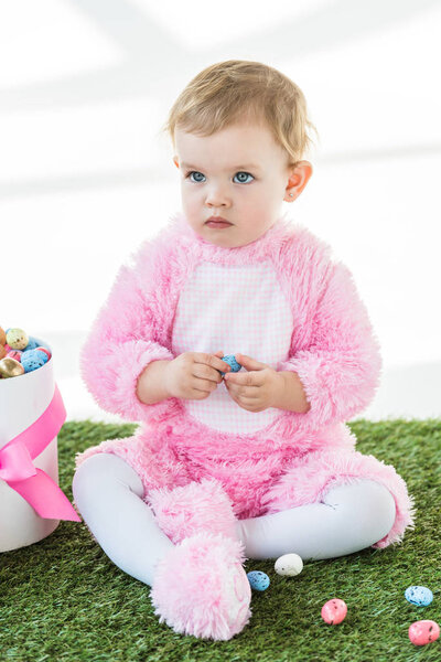 adorable child in pink fluffy costume holding blue quail egg while sitting near box with Easter eggs isolated on white