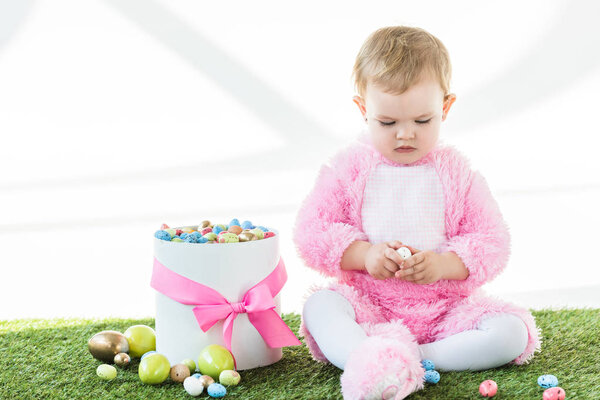 cute baby in pink fluffy costume holding quail egg while sitting near box with colorful Easter eggs isolated on white