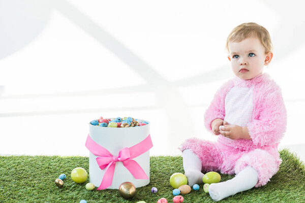 cute baby in pink fluffy costume sitting near box with colorful Easter eggs isolated on white