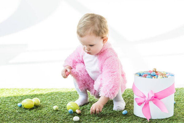 очаровательный ребенок в розовом пушистом костюме берет красочные яйца из зеленой травы возле подарочной коробки с розовым луком изолированы на белом
