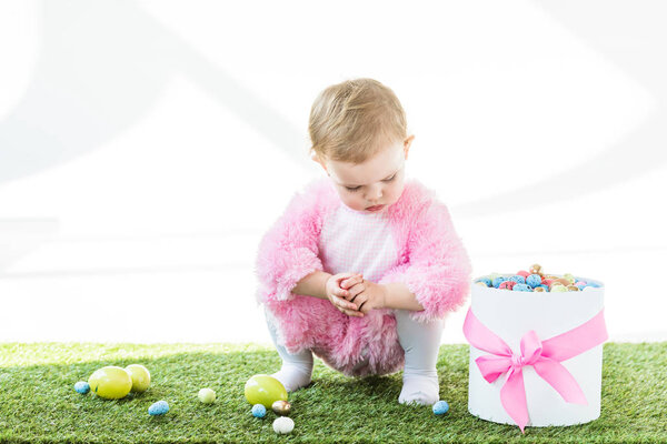 cute baby in pink fluffy costume sitting on green grass near gift box with pink bow and colorful Easter eggs isolated on white