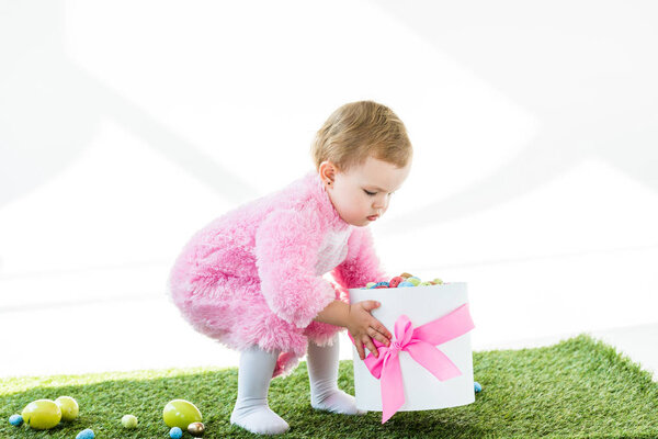 очаровательный ребенок в розовом пушистом костюме держит подарочную коробку с розовым луком и красочные пасхальные яйца изолированы на белом
