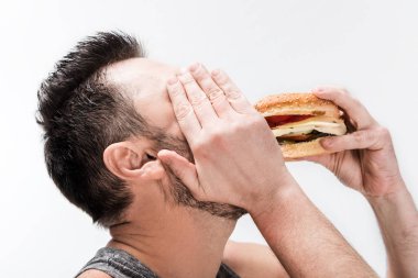 chubby bearded man covering face with hand while eating burger isolated on white clipart