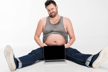 skeptical overweight man looking at camera while sitting near laptop with blank screen on white clipart