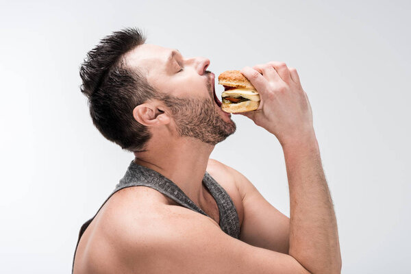 side view of chubby bearded man eating delicious burger isolated on white