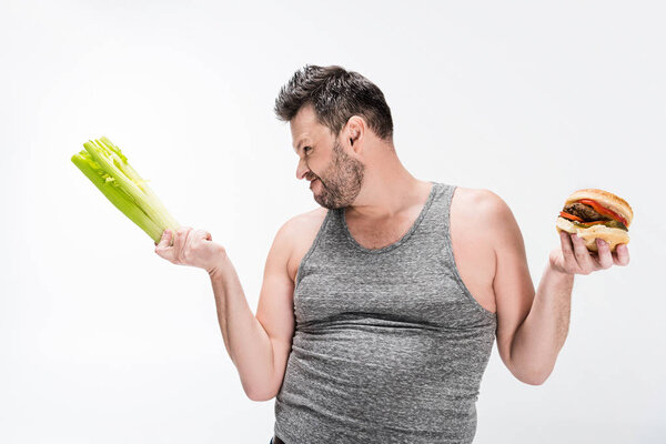 skeptical overweight man holding celery and burger isolated on white 