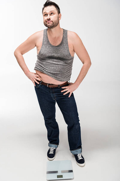 задумчивый человек с избыточным весом позирует с руками акимбо рядом с электронными весами, изолированными на белом
