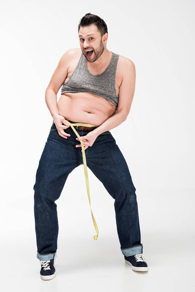 excited overweight man looking at camera and measuring waistline with tape on white