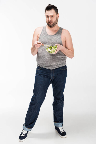 dissatisfied overweight man looking at camera and holding bowl of salad on white