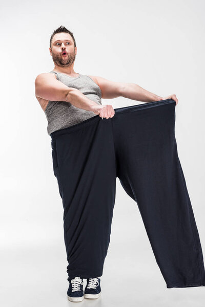 surprised overweight man holding oversize pants after weight loss on white