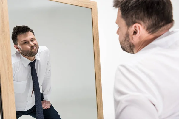 chubby man in formal wear looking at reflection in mirror isolated on white