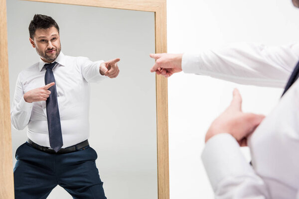 overweight man in formal wear pointing with fingers while looking at mirror isolated on white