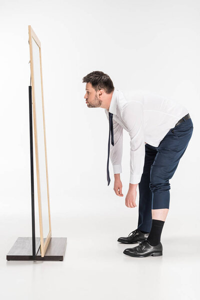 толстый мужчина в формальной одежде наклоняется и смотрит на зеркало на белом
