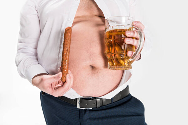 partial view of overweight man showing belly and holding glass of beer with grilled sausage isolated on white