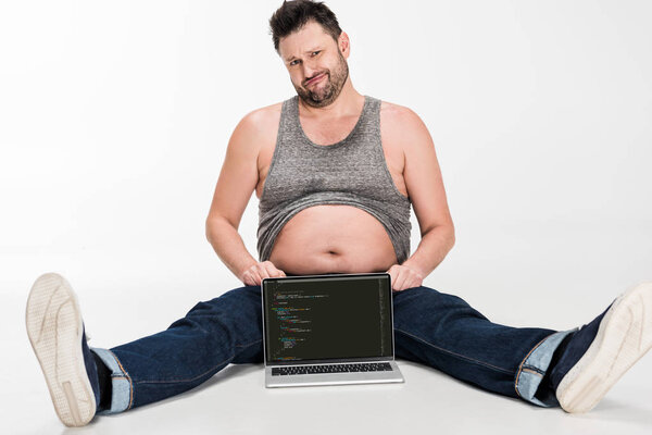 skeptical overweight man making facial expression and sitting with laptop with microsoft windows software on screen isolated on white