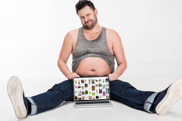 skeptical overweight man making facial expression and sitting with laptop with pinterest website on screen isolated on white