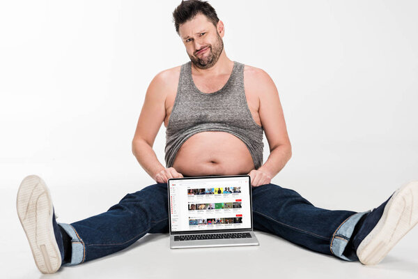 skeptical overweight man making facial expression and sitting with laptop with youtube website on screen isolated on white