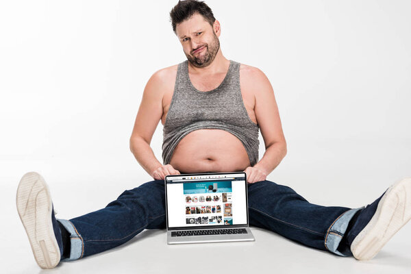 skeptical overweight man making facial expression and sitting with laptop with amazon website on screen isolated on white