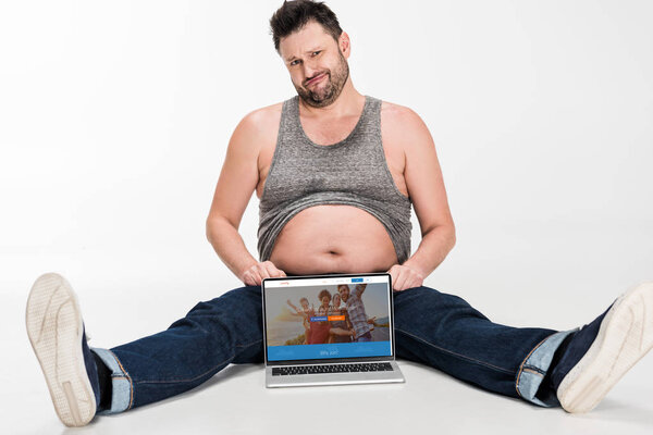 skeptical overweight man making facial expression and sitting with laptop with couchsurfing website on screen isolated on white