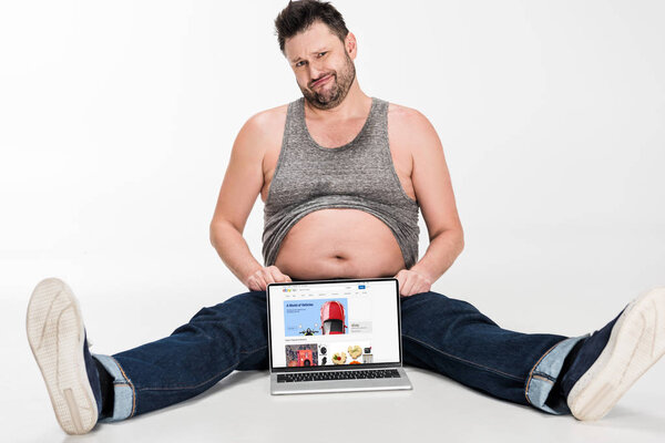 skeptical overweight man making facial expression and sitting with laptop with ebay website on screen isolated on white