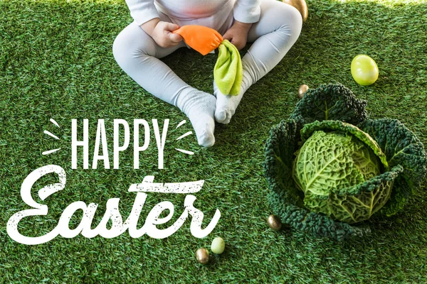 儿童拿着玩具胡萝卜 而坐在复活节彩蛋和卷心菜在绿草地上与快乐复活节字母的部分视图 — 图库照片