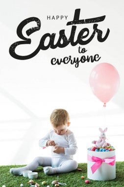 cute baby sitting near box with colorful quail eggs, toy rabbit and air balloon on white background with happy Easter to everyone lettering  clipart