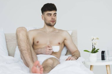 selective focus of handsome, thoughtful man in underpants lying in bed with cup of coffee clipart