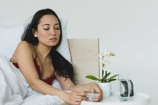 beautiful, sad asian woman holding birth control pills while lying in bed near table with orchid in flowerpot