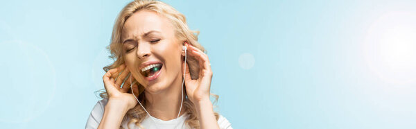 panoramic shot of cheerful woman with closed eyes listening music in earphones and singing on blue 
