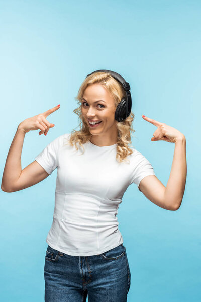 счастливая блондинка, указывающая пальцами на черные наушники на синем
 